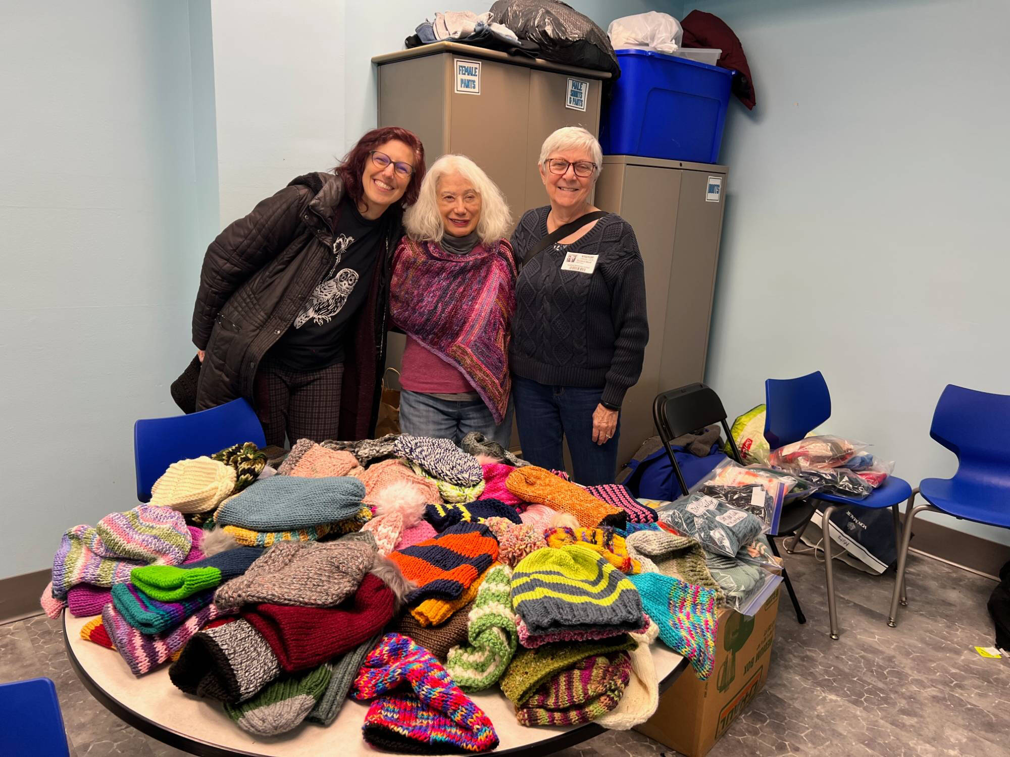 Three women behind a large table full of colorful knitted hats, scarves, and mittens.